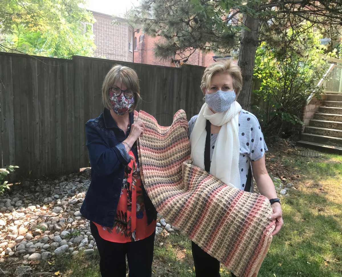 Giselle Weaver (L) giving one of six blankets to Melodie Pritchard (R). Although their smiles are concealed, the gift of loving care speaks loudly as they hold one of seven lap blankets made by Giselle. 